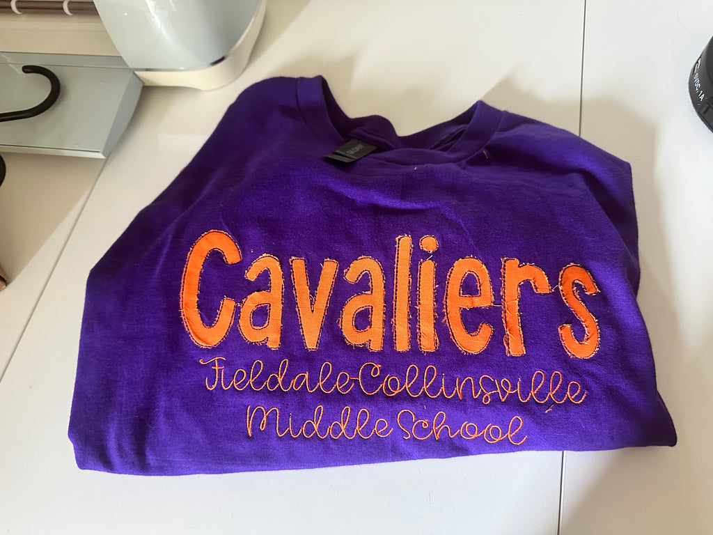 Purple shirt with 'Cavaliers' and school name printed on it, laid flat on a surface.